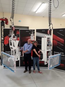 Two Binar Handling SAS employees with big smiles holding two Ergo Glass end effectors in front of four Binar Handling roll-ups behind them